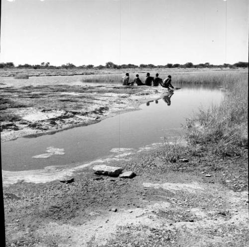 Five men sitting on the sand at the edge of a pan, distant view from behind