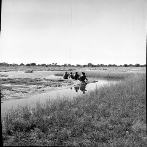 Five men sitting on the sand at the edge of a pan, distant view from behind