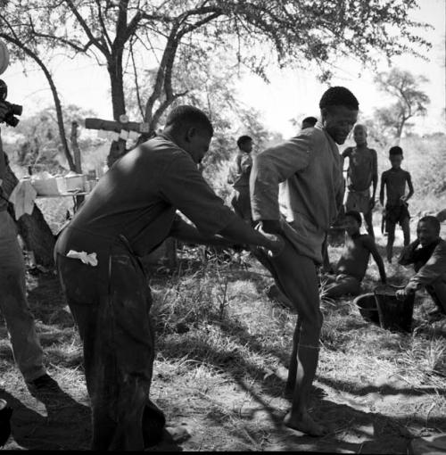 Man receiving a shot from the expedition doctor, with a group of people standing in the background