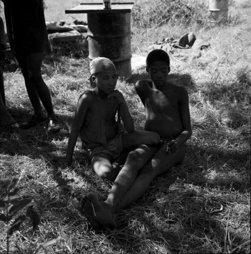 Two boys sitting, with metal drums in the background