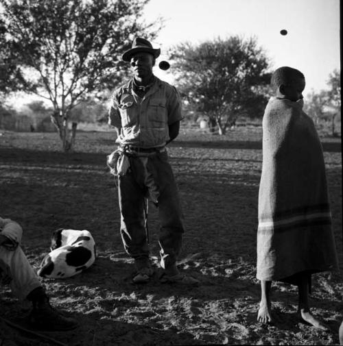 Man wrapped in a blanket standing next to a man wearing Western clothing, with a dog lying on the ground near them