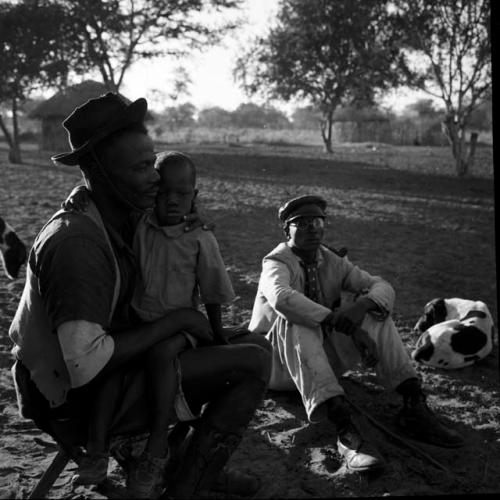 Man sitting on a stool, holding a child, with a man wearing a white suit sitting on the ground next to him, smoking a pipe, dog lying on the ground next to him