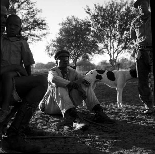 Man sitting on a stool, holding a child, with a man wearing a white suit sitting on the ground next to him, smoking a pipe, a man and a dog standing next to him