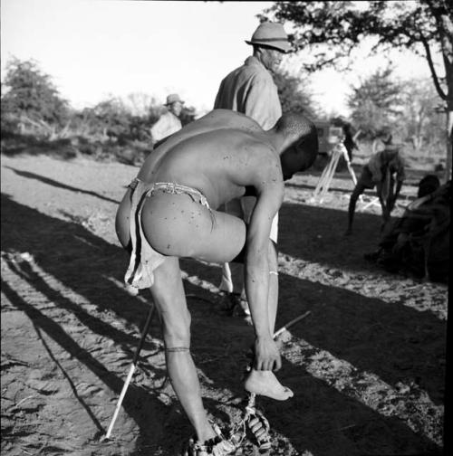 Man leaning over to put his dance rattles on, view from behind, with /Ti!kay standing behind him, camera on a tripod in the background