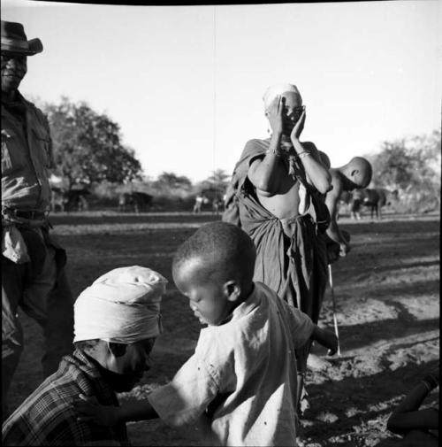 Woman standing, holding her hands to the sides of her face, with a man wearing western clothing and a man putting on dance rattles standing next to her, woman with a child sitting near them