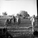 /Ti!kay standing (right), with people sitting and standing near him, cattle in the background