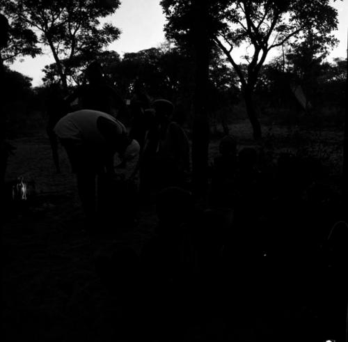 Tsamgao wearing a white T-shirt, leaning over a jerry can in front of a fire, with people sitting and standing near him