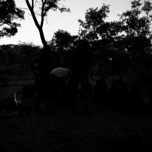 Tsamgao wearing a white T-shirt, leaning over a jerry can in front of a fire, with people sitting and standing near him