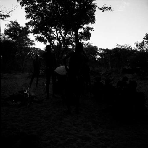 Tsamgao wearing a white T-shirt, leaning over a jerry can in front of a fire, with people sitting and standing near him