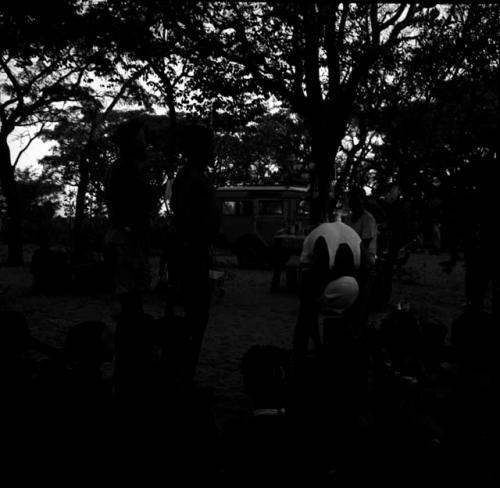 Tsamgao wearing a white T-shirt, leaning over a jerry can in front of a fire, with the expedition Land Rover in the background