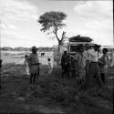Group of men and children standing next to the expedition Land Rover, with a donkey in the background, tree in the distance