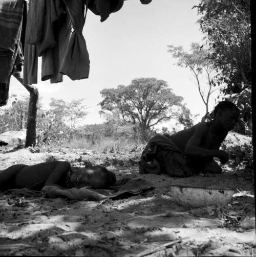 Woman kneeling next to a sleeping child, with blankets hanging near them, enamel dish on the ground