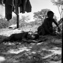 Woman kneeling next to a sleeping child, with blankets hanging near them, enamel dish on the ground
