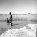 Man walks through water in a pan, with a man sitting in the water, bathing behind him