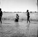 Man standing in the water in a pan, with John Marshall squatting in the water, filming him