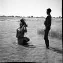 Man standing in the water in a pan, with John Marshall kneeling in the water, filming him