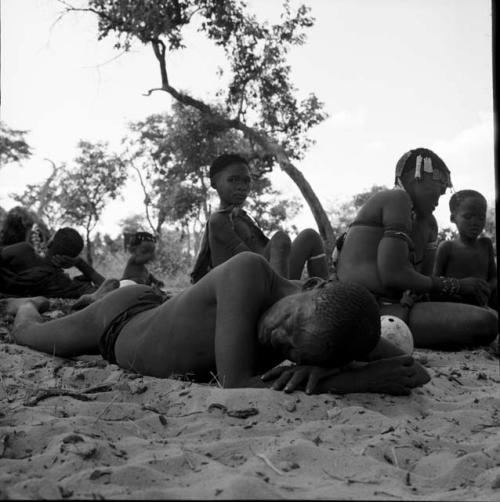 Man lying on the ground, sleeping, with a woman wearing ornaments in her hair sitting with his family behind him