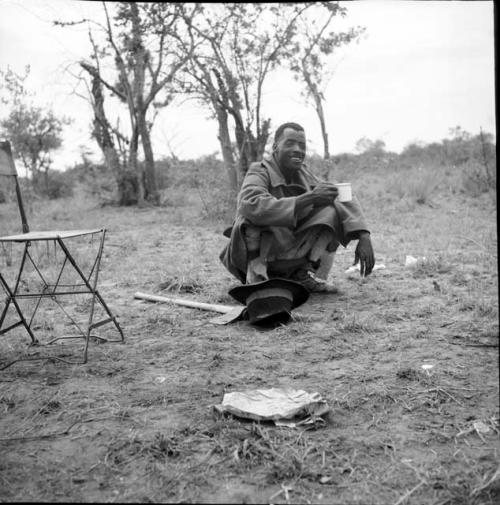 Man from Kubi squatting, drinking from an enamel cup, with his hat and his axe on the ground next to him