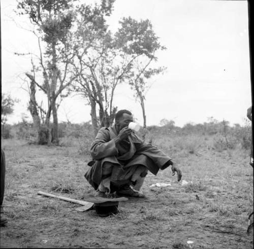 Man from Kubi squatting, drinking from an enamel cup, with his hat and his axe on the ground next to him