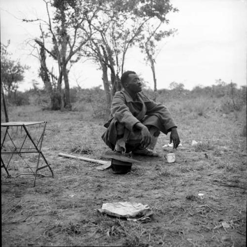 Man from Kubi squatting, with an enamel cup, his hat and his axe on the ground next to him