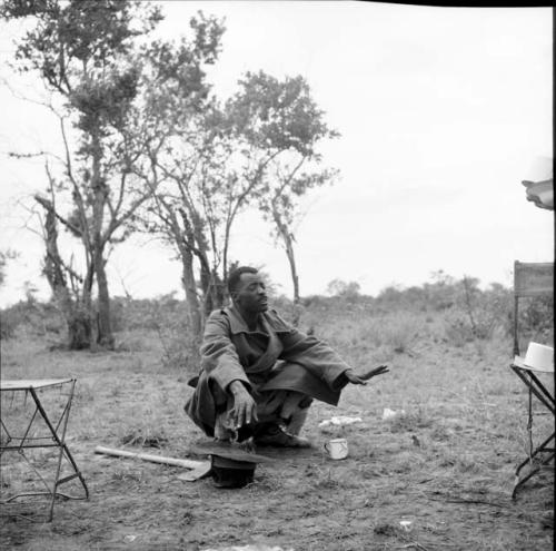 Man from Kubi squatting, gesturing with his hands, with an enamel cup, his hat and his axe on the ground next to him