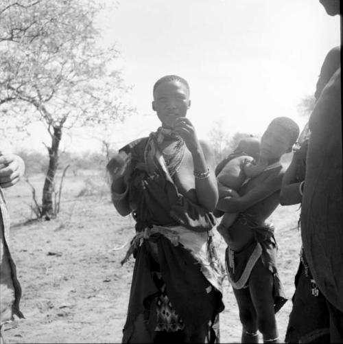 Woman wearing a furred animal skin around her waist, standing, talking, with a girl holding a baby standing next to her, listening