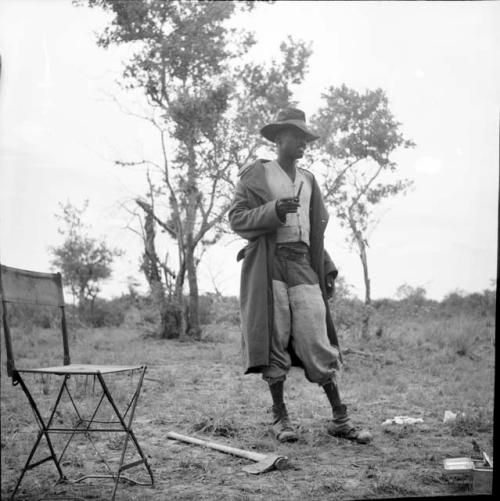 Man from Kubi standing, smoking a pipe, with his axe and an expedition chair on the ground next to him