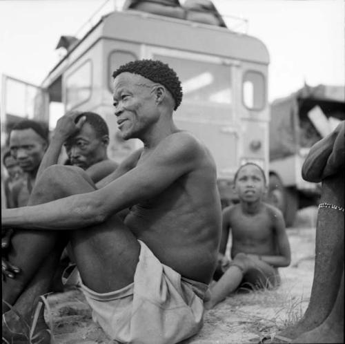 /Ti!kay and Bojo sit with other men and a boy in front of the expedition Land Rover