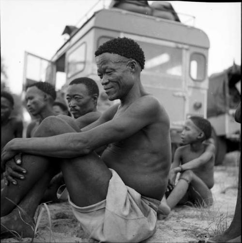 /Ti!kay and Bojo sit with other men and a boy in front of the expedition Land Rover