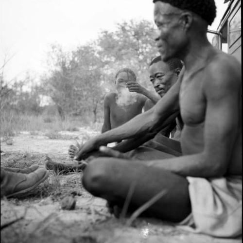 /Ti!kay and Bojo sitting with another man in front of the expedition Land Rover