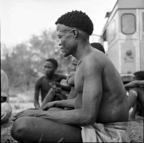 /Ti!kay sitting with other men in front of the expedition Land Rover