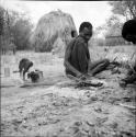 Man heating an arrow point in a fire, with a puppy sniffing the ground next to him