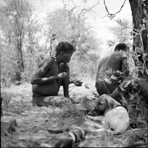 Man holding arrows in his hand, checking them for sharpness, sitting with two other men and a puppy