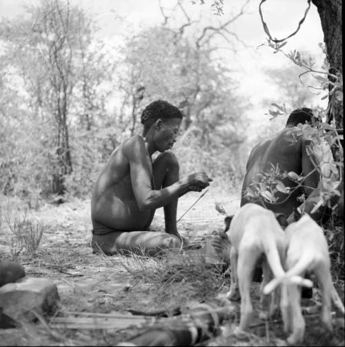 Man working on an arrow point, sitting with another man, with two puppies standing next to them