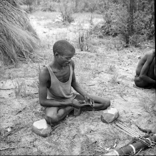 "/Gao Music" wearing an undershirt, sitting, testing an arrow, with a quiver and arrows on the ground next to him