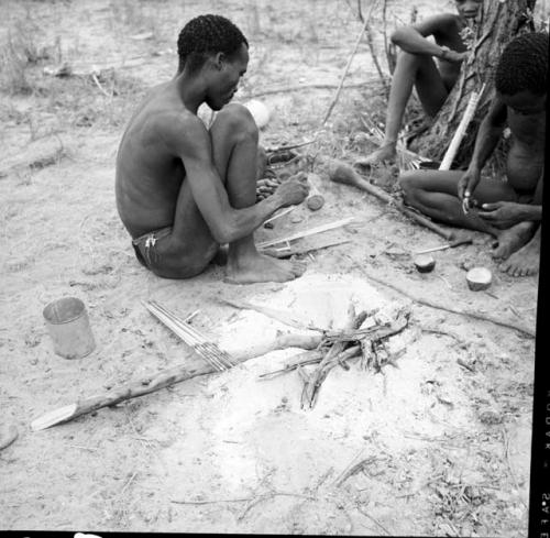 Bojo sharpening an arrow point with a stone, with arrows resting on a stick, ostrich eggshell and dish of poison grubs on the ground near him