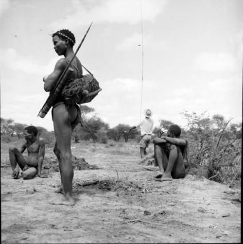 Man standing, carrying hunting equipment, view from behind, with two men sitting behind him, man wearing Western clothing adjusting a spring hare pole in the background