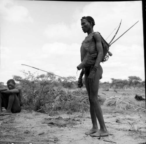 Man standing, carrying hunting equipment, holding a spring hare pole, with a man sitting behind him