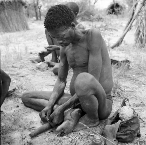 Man working on an arrow, with a piece of veldkos and a sock in a bag on the ground next to him