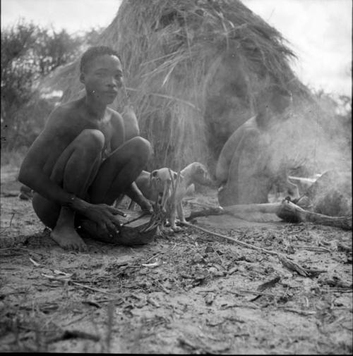 Bojo eating, sitting in front of his skerm next to a man playing a //guashi, with dogs standing next to them