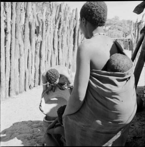 Woman with a baby on her back in her kaross standing, watching another woman pour corn from a large basin into a small bucket