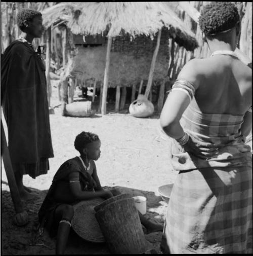 Woman sitting, working with corn in a hollow log, with two women standing next to her, with a skerm in the background