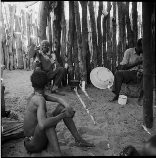 Group of people sitting inside a skerm, with an enamel basin leaning against a fence near them