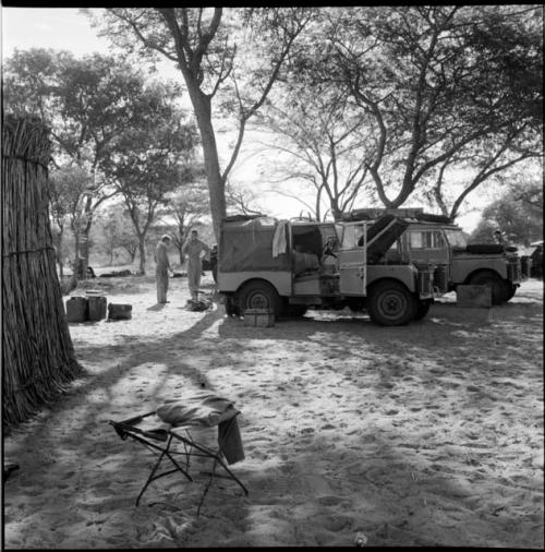 Two expedition members stand next to the Land Rover and the Jeep, with luggage and a stool on the ground near them