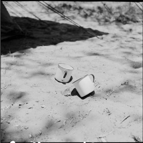 Two enamel cups in the sand next to the shadow of an expedition tent