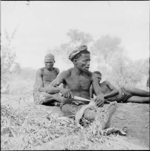 ≠Toma wearing a skin hat, cutting strips of skin with a large knife, with two men behind him