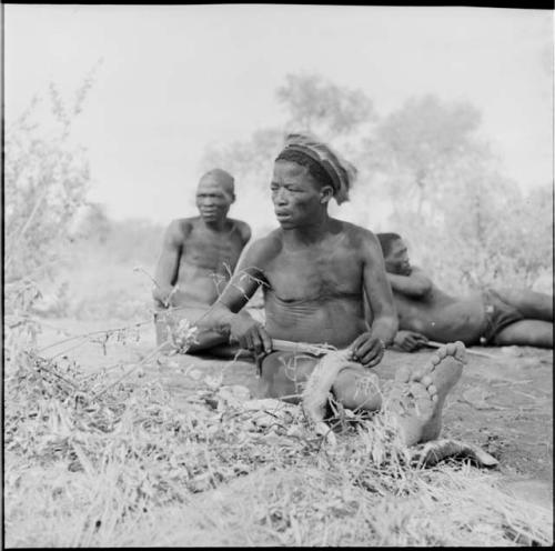 ≠Toma wearing a skin hat, cutting strips of skin with a large knife, with two men behind him