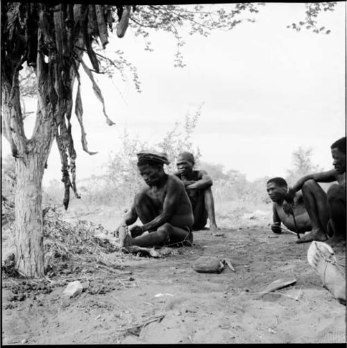 ≠Toma wearing a skin hat, cutting strips of skin with a large knife, with a group of men sitting behind him