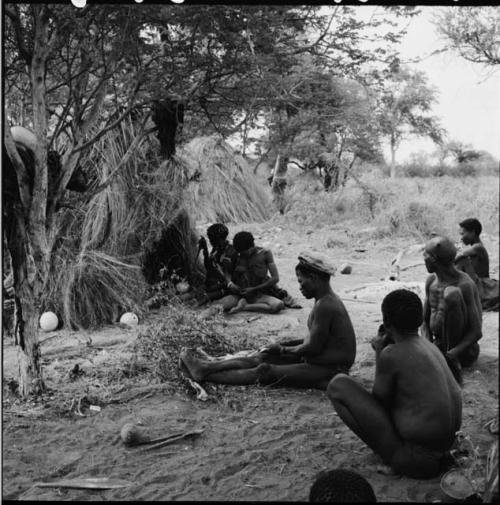 ≠Toma wearing a skin hat, cutting strips of skin with a large knife, sitting with a group of people near a skerm