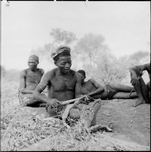 ≠Toma wearing a skin hat, cutting strips of skin with a large knife, with three men behind him